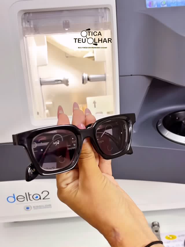 Mão segurando um óculos de sol de acetato preto com lente de grau, diante da máquina de montagem de lentes da loja.